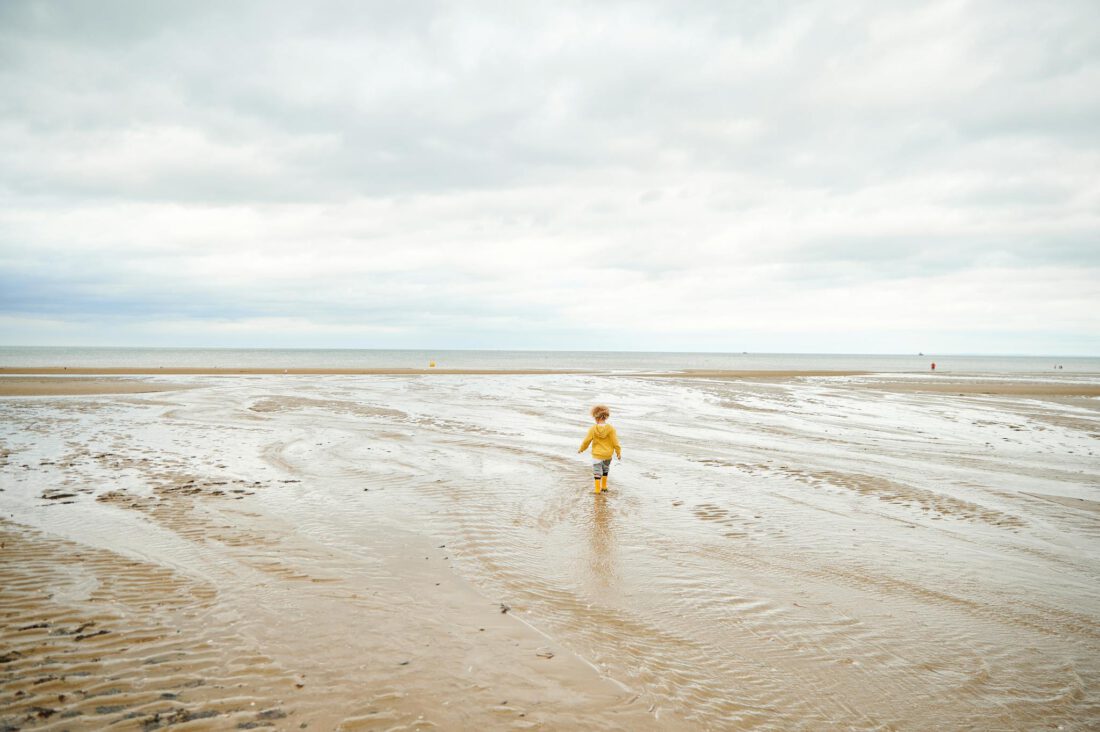 A child in yellow walks alone on a vast, sandy beach under a cloudy sky.