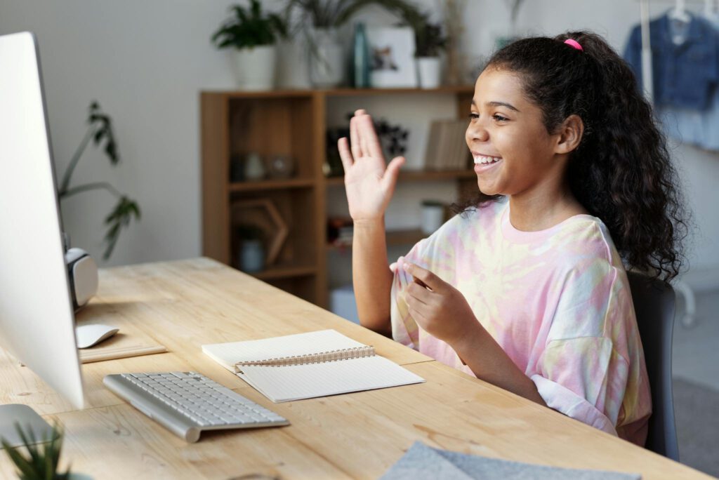 A happy girl waves during an online class at home, sitting at a desk with a computer.