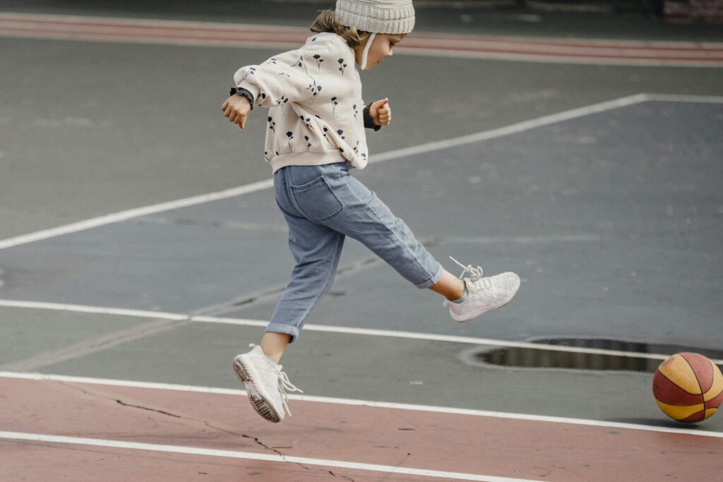 Side view of jumping little girl in jeans and jumper kicking colorful ball playing on sports yard